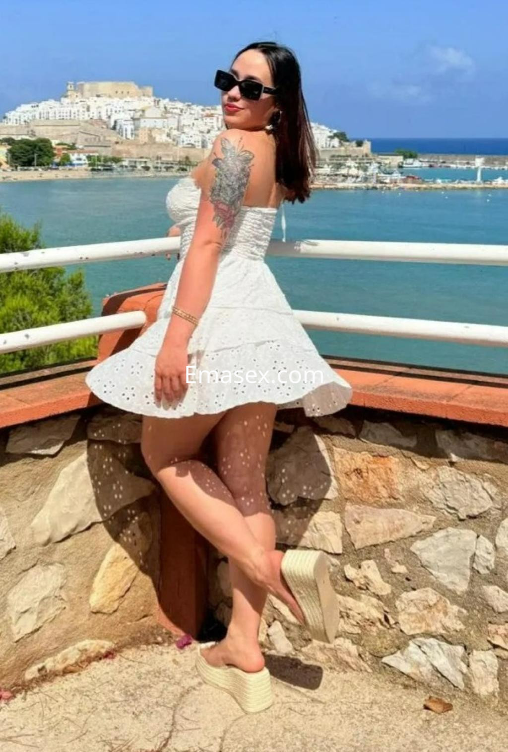 Escort en Alicante luciendo un vestido blanco corto y gafas de sol, posando con un paisaje costero de fondo.
