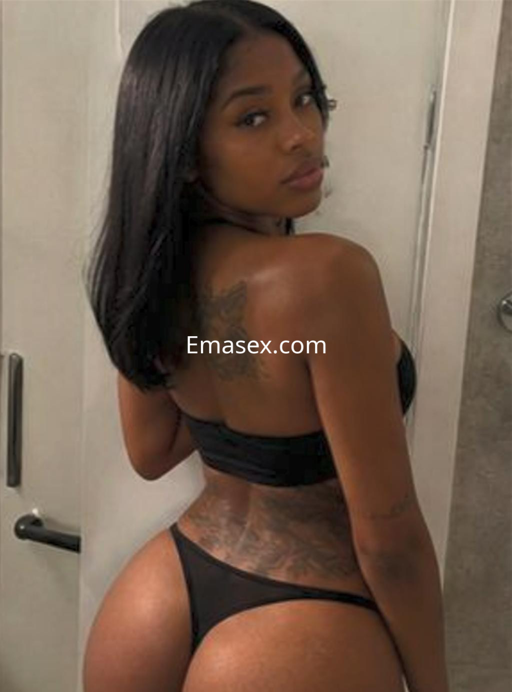 Escort en Vitoria con cabello lacio y oscuro, vistiendo un sujetador negro y braguitas, mostrando un tatuaje en la espalda.