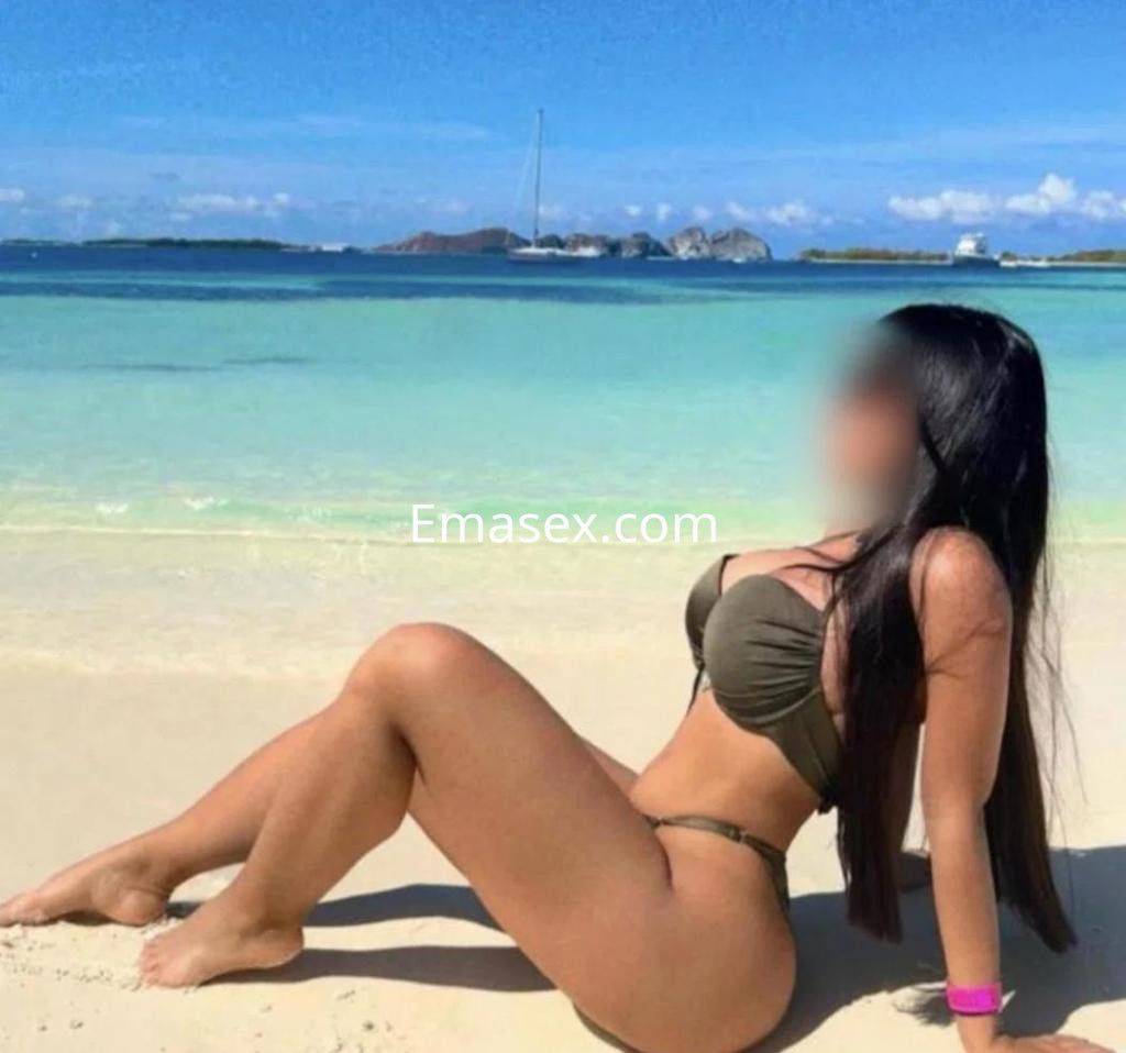 Escort en Cádiz luciendo un bikini verde, sentada en la playa con el mar y un velero de fondo.
