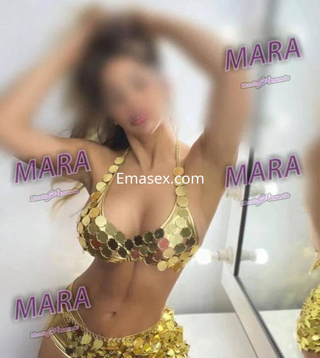 Escort en Cádiz con top dorado brillante de monedas, posando con una expresión coqueta y un fondo neutro.