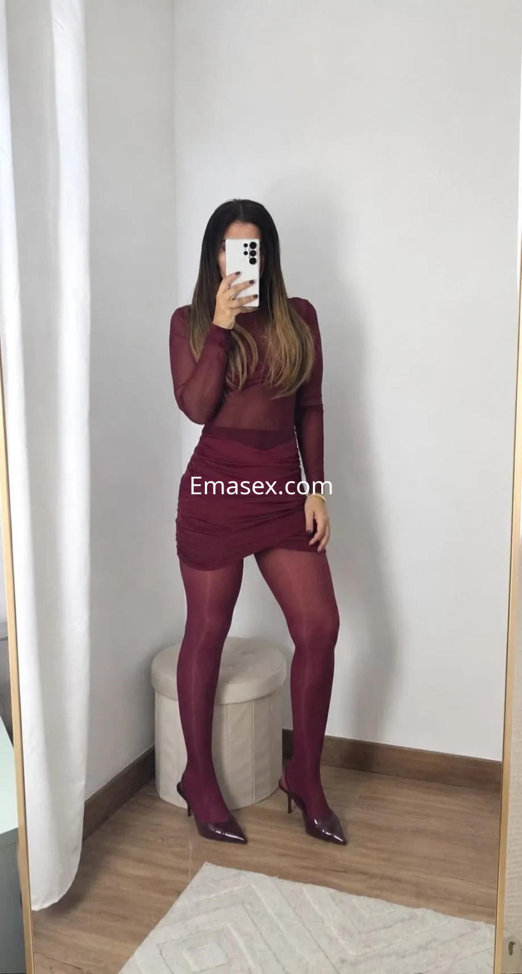 Escort en Coruña con un vestido ajustado de color burdeos y pantys a juego, posando frente al espejo.