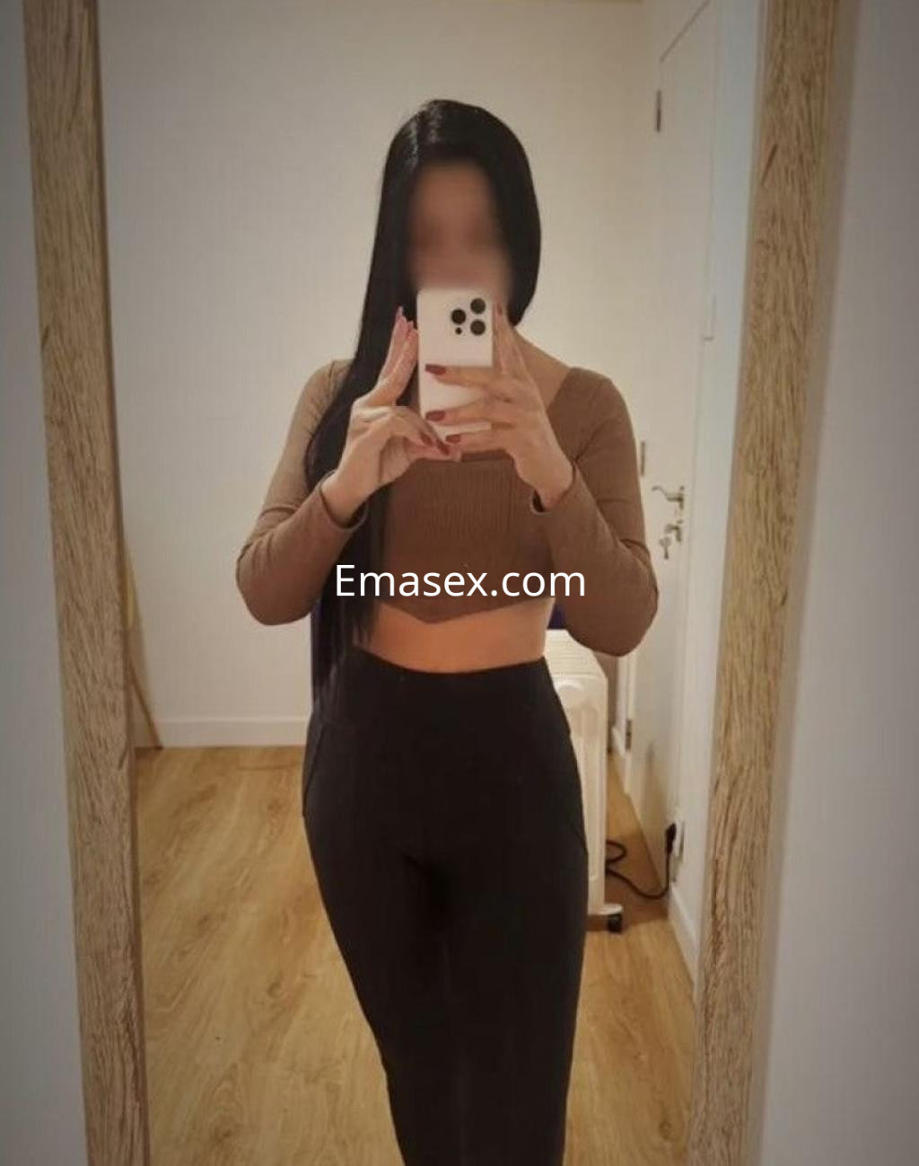Escort en Coruña, posando frente a un espejo con top de manga larga marrón y pantalones negros ajustados.