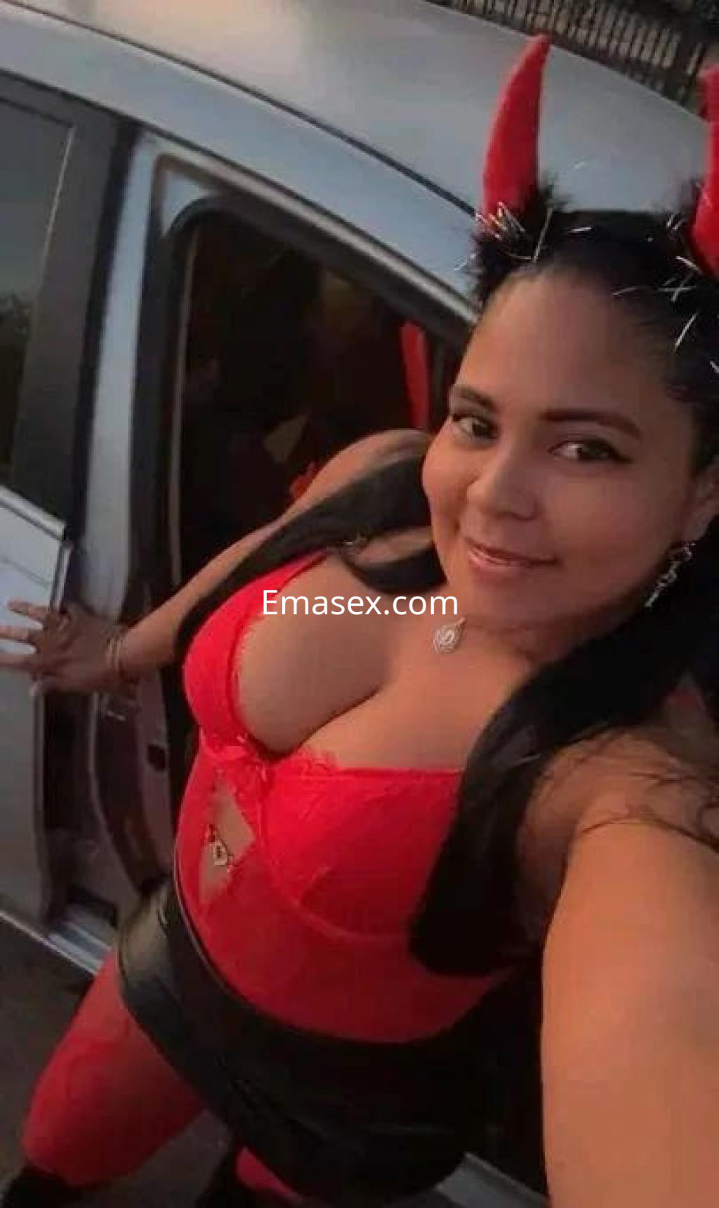 Escort en Huelva, posando con una blusa roja y una falda negra, junto a un automóvil.