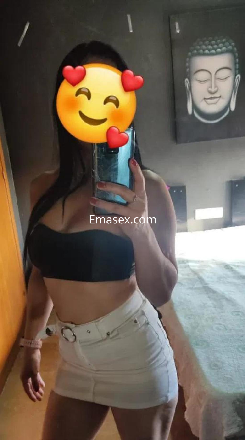 Escort en León posa frente a un espejo, luciendo un top negro y una falda corta blanca, con fondo decorativo.