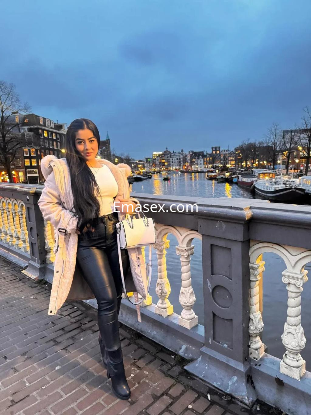 Escort en Murcia posando junto al canal, viste abrigo claro y leggings negros, con cabello largo y oscuro.