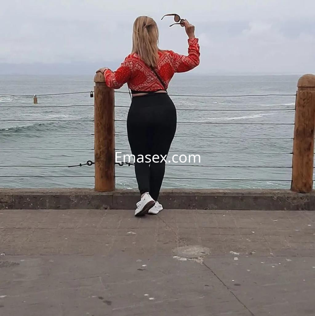 Puta con cabello rubio y lacio, viste blusa roja y pantalones ajustados, posando de espaldas en la costa de Murcia.