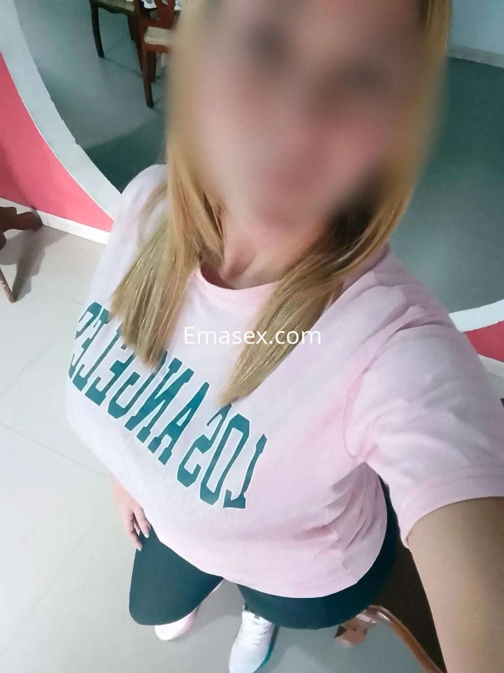 Puta de cabello rubio, con camiseta rosa y pantalones deportivos, posando en un ambiente interior en Gijón.