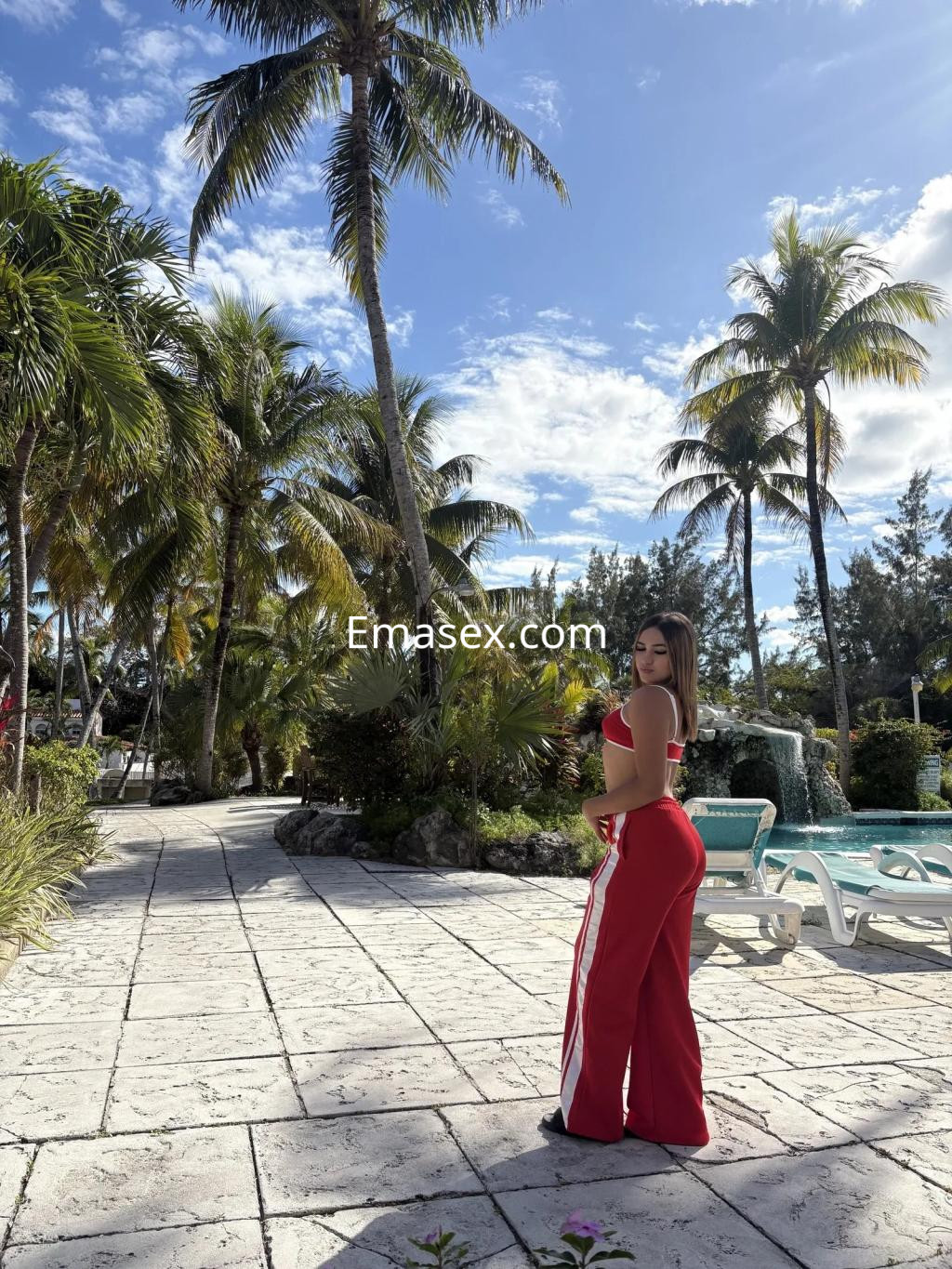 Escort en Vigo, luciendo un conjunto rojo y posando al aire libre entre palmeras y una piscina.