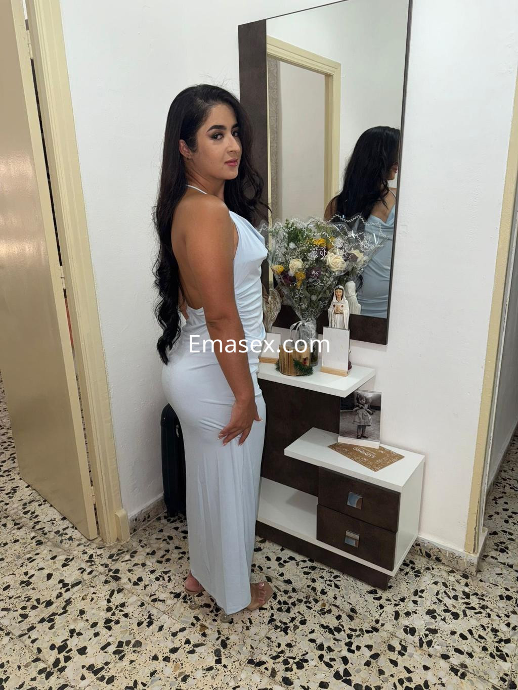 La puta en Vigo viste un elegante vestido azul claro y posa frente a un espejo, rodeada de flores.