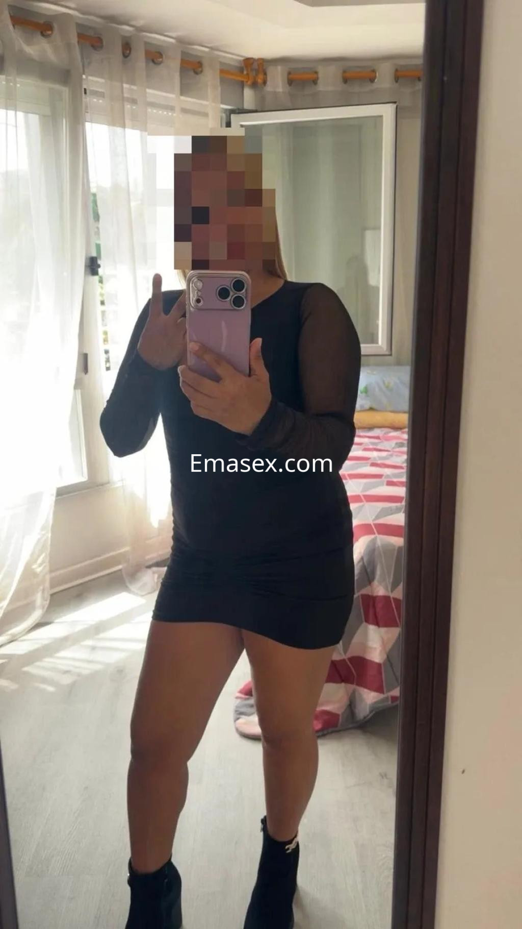 La puta en Vigo se muestra en un vestido negro ajustado y botines, posando frente a un espejo.