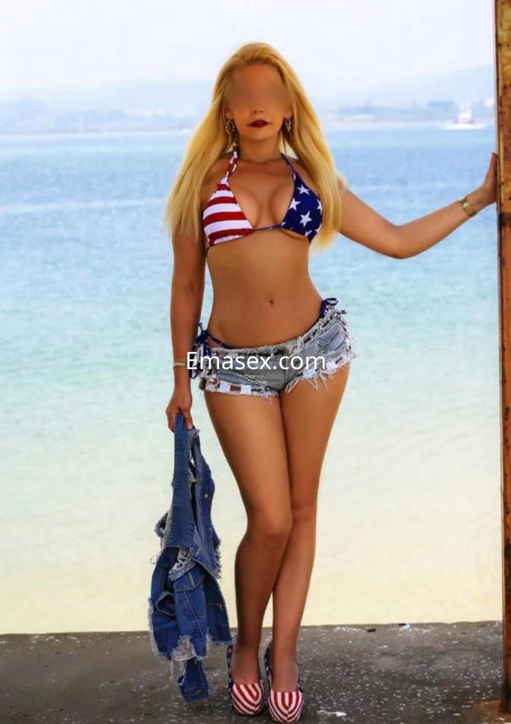 Puta en Salamanca con bikini de banderas, short de mezclilla y sandalias, posando junto al mar.