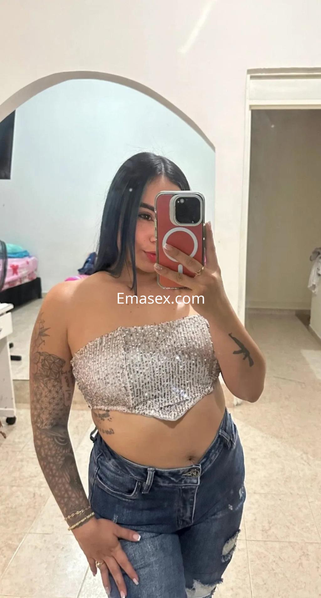 Escort en Sevilla posando frente a un espejo, con top brillante y jeans ajustados, mostrando tatuajes en su brazo.