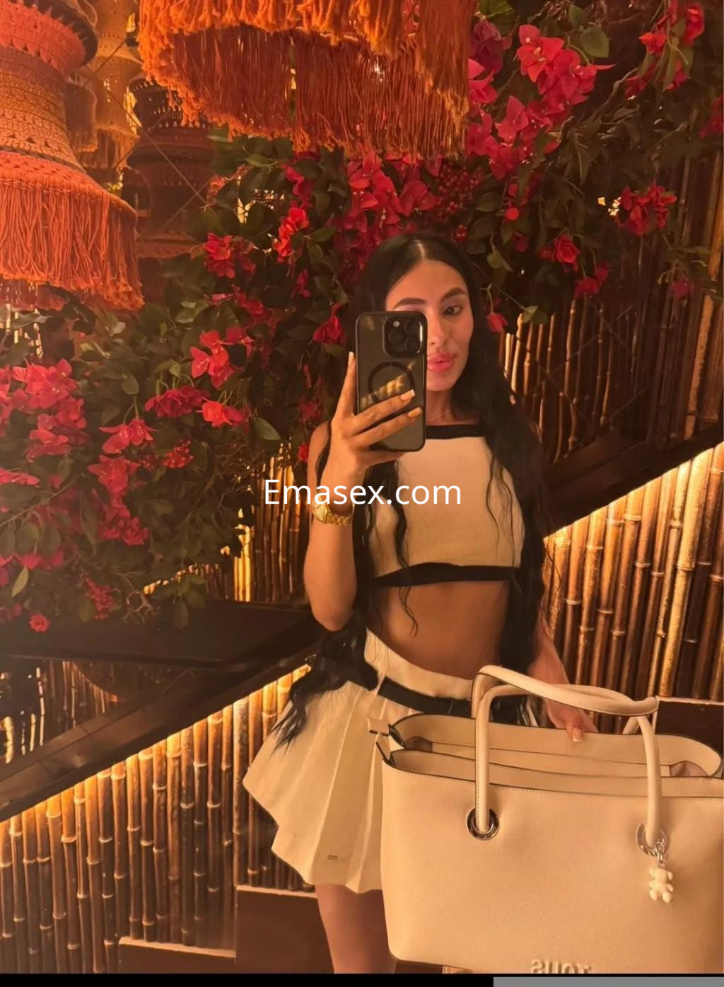 La puta en Toledo posa con un top negro y una falda blanca, sosteniendo un bolso claro en un entorno decorado con flores.