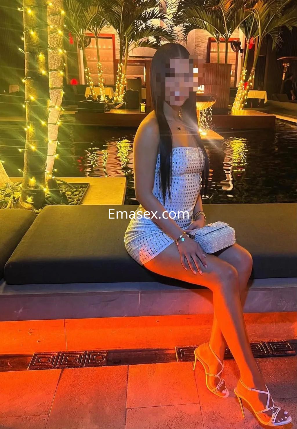 Escort sentada en un sillón junto a una piscina iluminada, lleva un vestido ajustado y tacones altos en Valladolid.