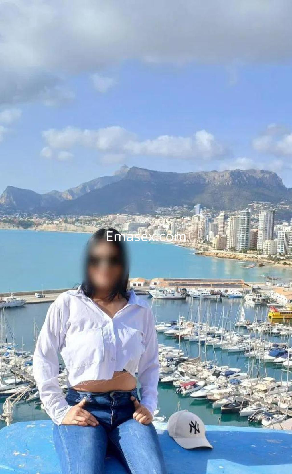 Escort posando en un mirador de Huelva, luciendo una camisa blanca y jeans, con un paisaje de puerto al fondo.