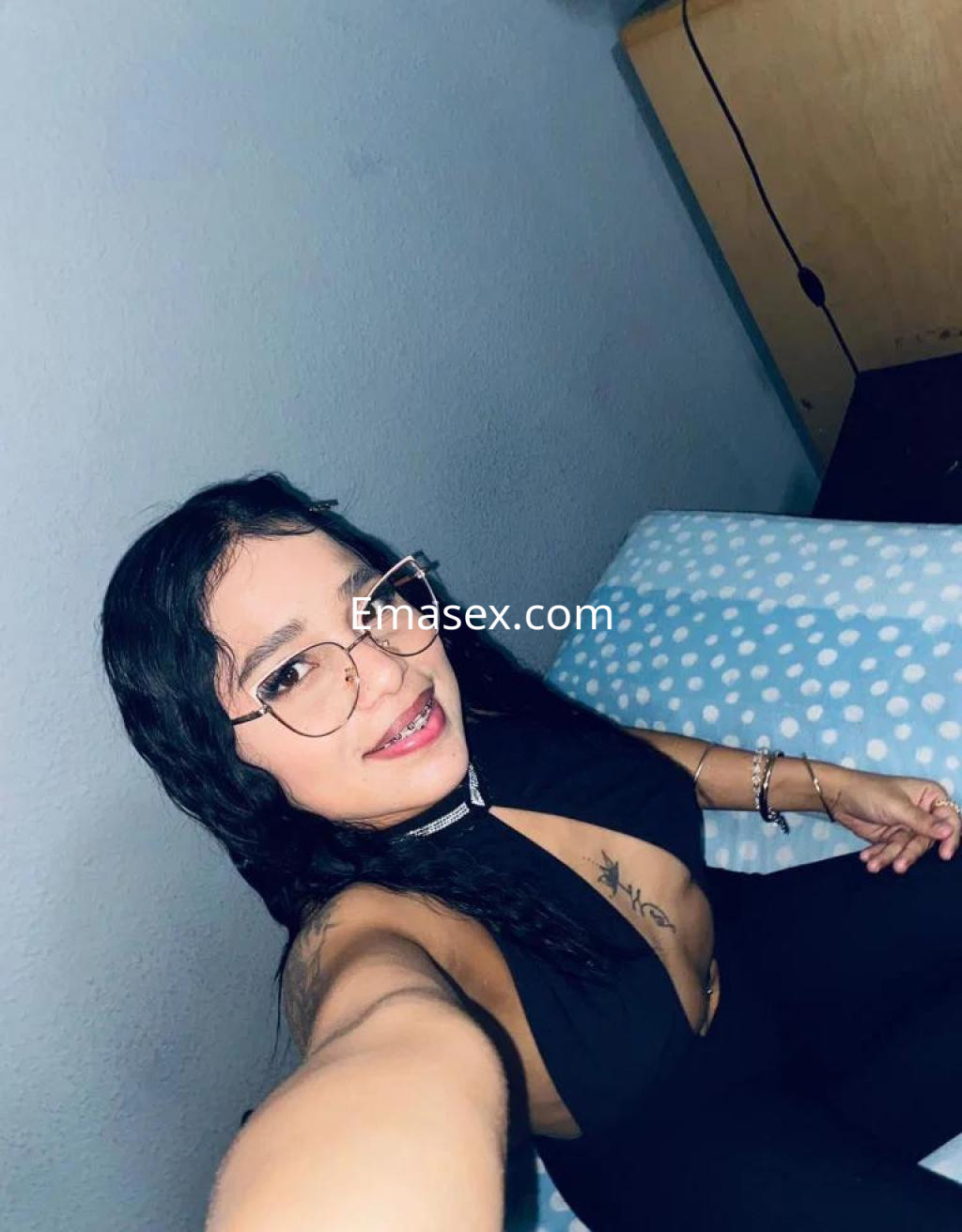 Escort en Salamanca con cabello oscuro y ondulado, usando gafas y un vestido negro, posando en una cama.