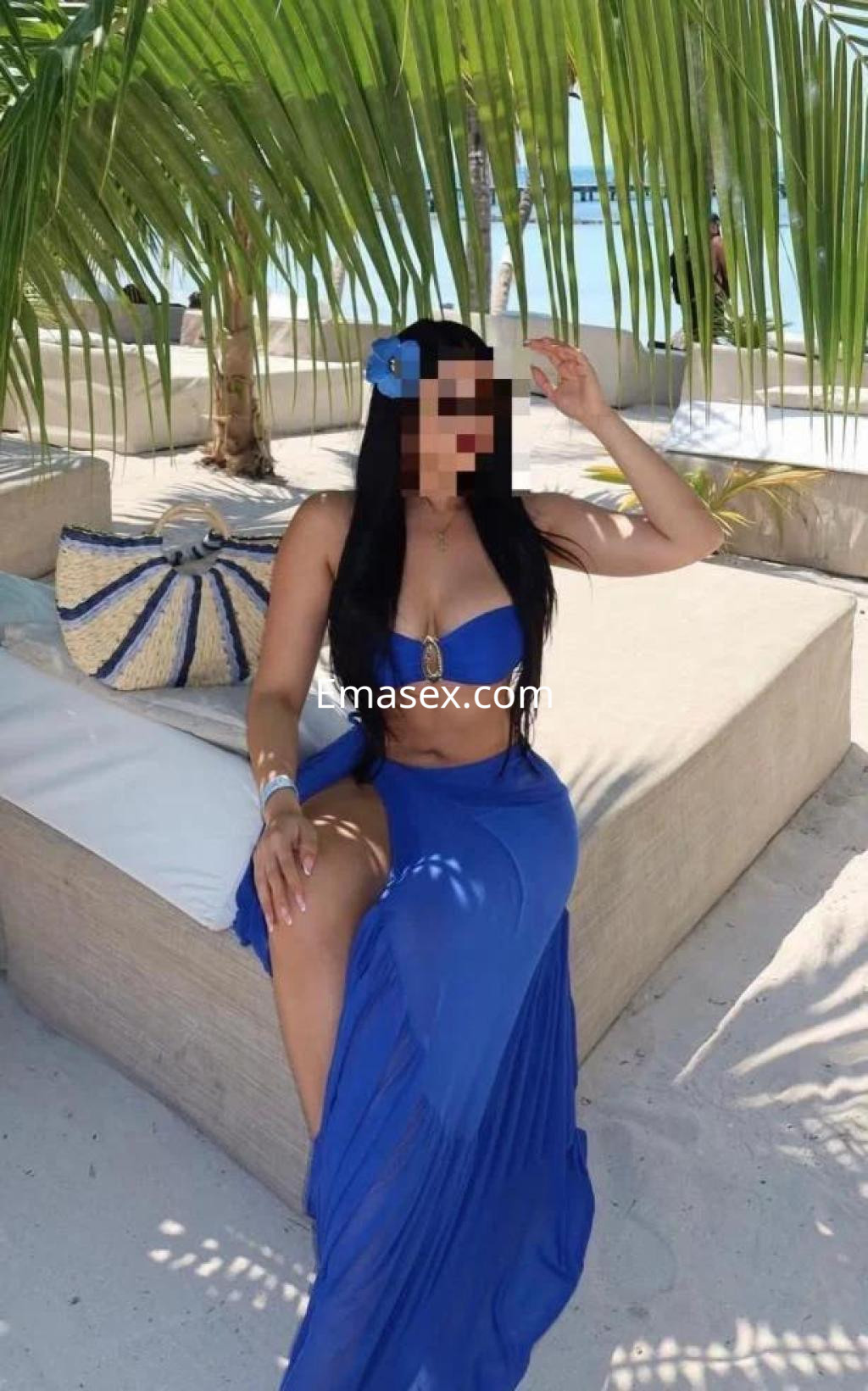 La puta posa en una playa de Albacete, luciendo un bikini azul y una falda larga, rodeada de palmeras.