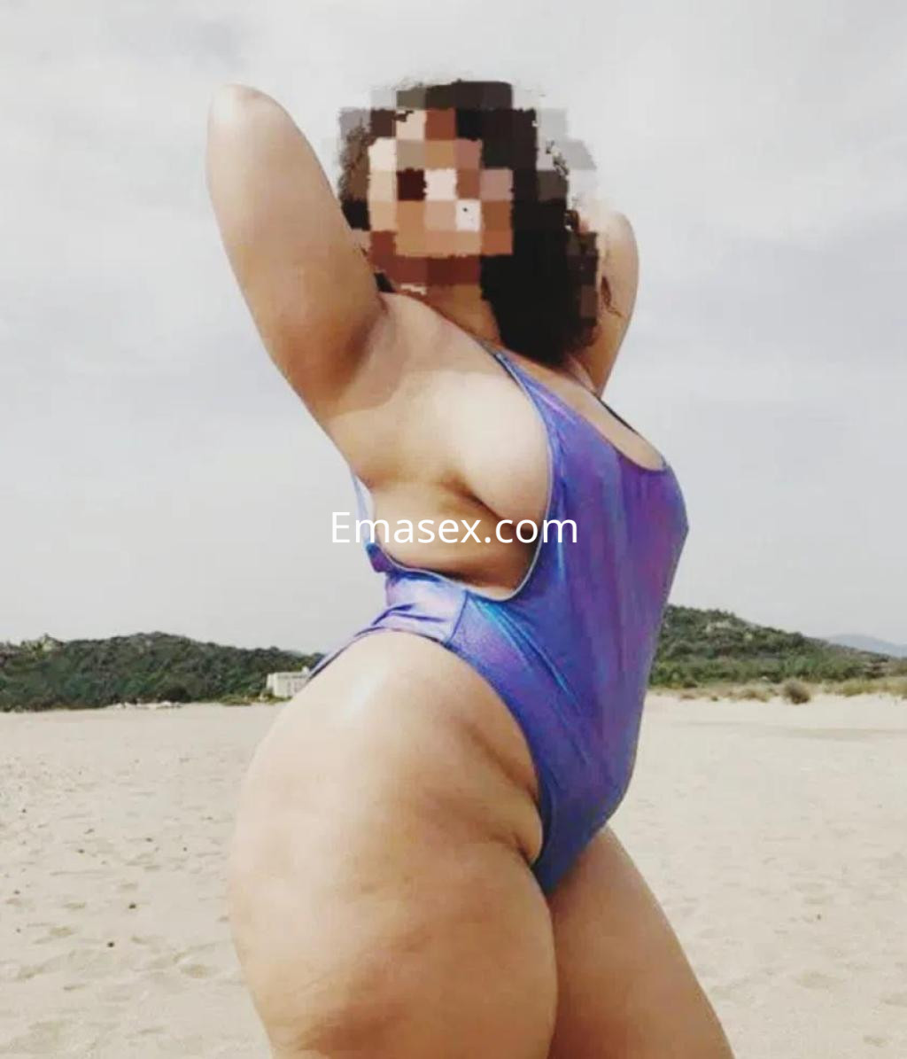 Escort posando en la playa de Palma de Mallorca, con un bañador de color morado y cabello rizado.