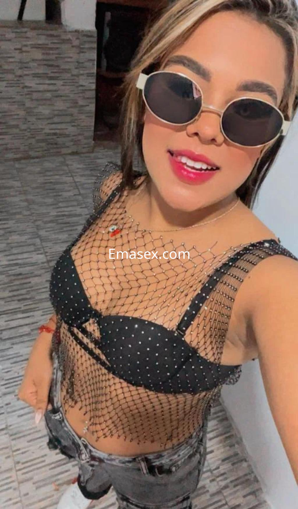 La puta en León viste una blusa de red con detalles en perlas y jeans, luciendo gafas de sol estilizadas.