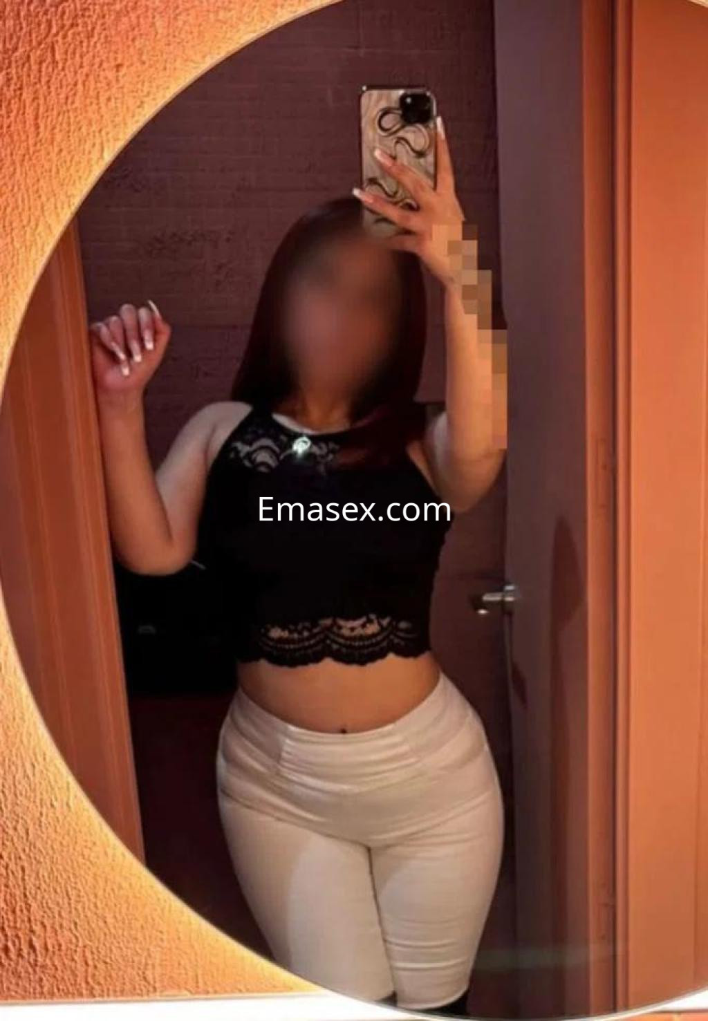 La puta en Madrid se refleja en un espejo, luciendo un top negro y pantalones blancos ajustados.
