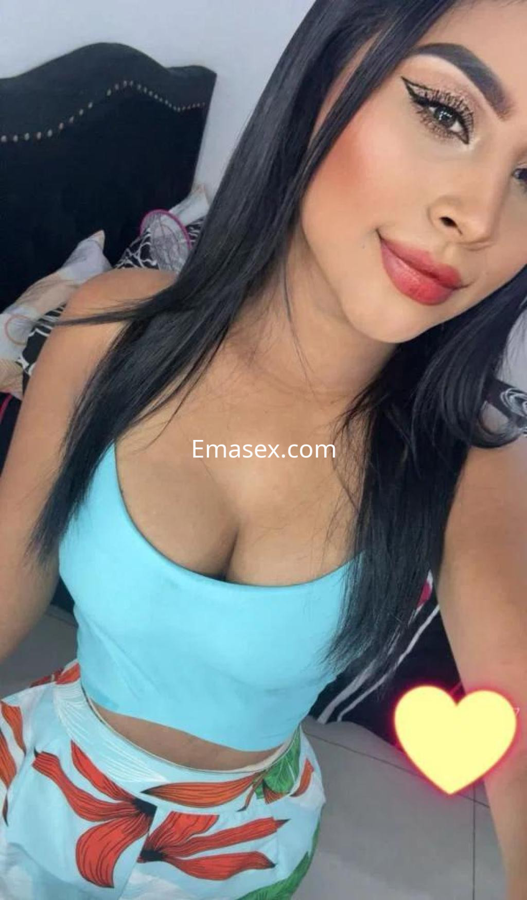 Escort en Madrid con cabello largo y lacio, vistiendo un top azul y una falda de flores, sonriendo en un entorno acogedor.