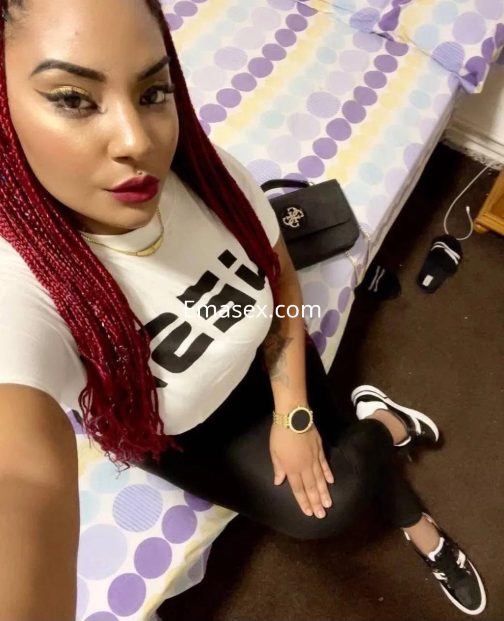 Puta con cabello largo y trenzado, viste camiseta blanca y leggings negros, sentada en una cama en Murcia.