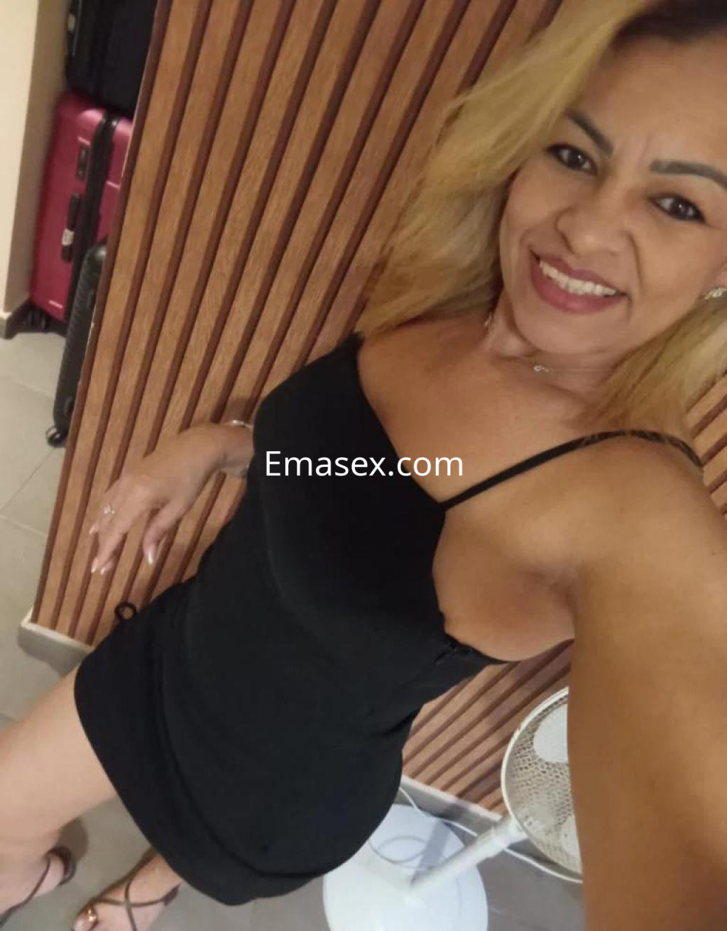 Puta en Oviedo luce un vestido negro ajustado y sandalias, sonriendo frente a una pared de madera.