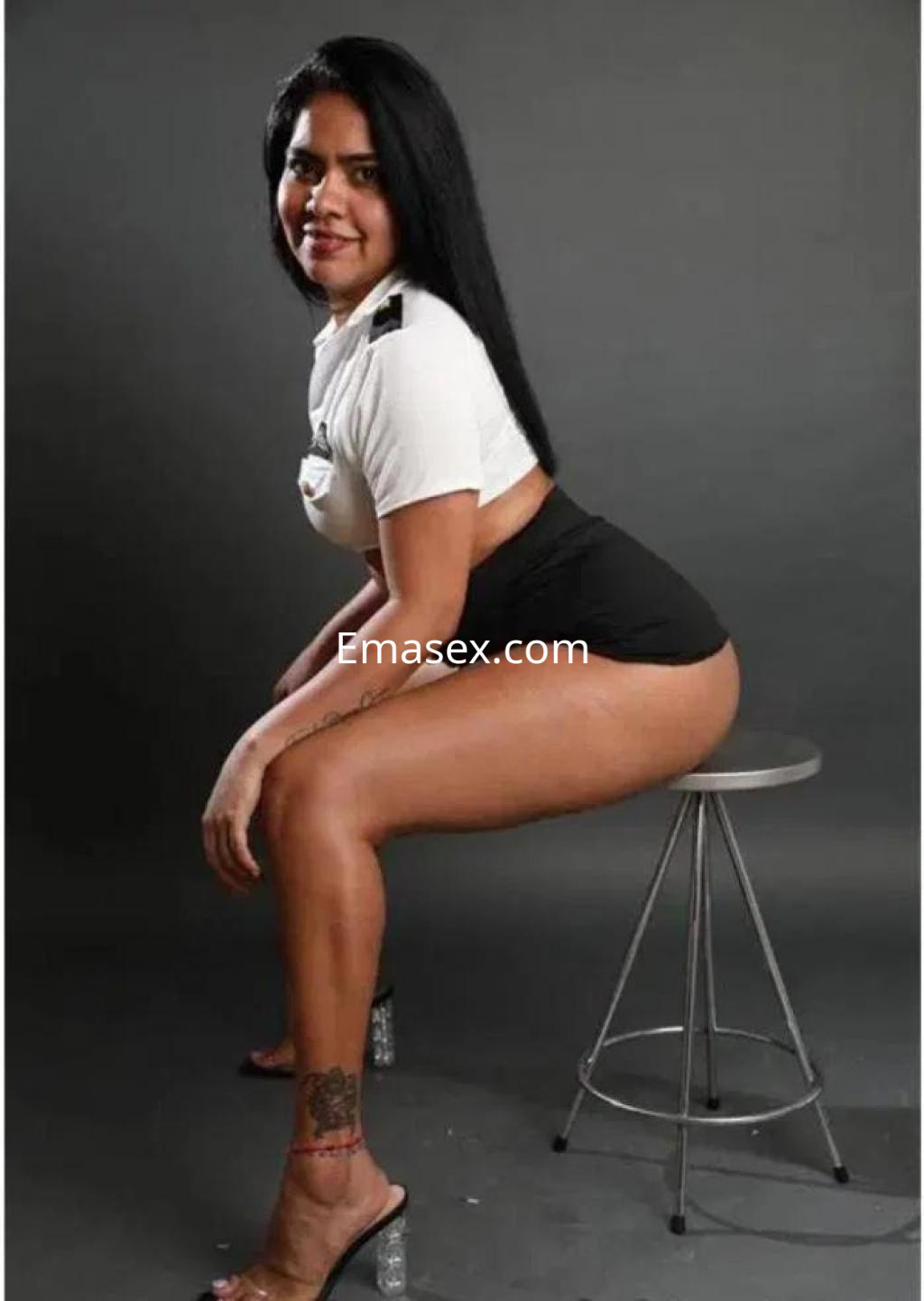 Puta de Salamanca posa sentada en un taburete, luciendo un top blanco y shorts negros, con cabello largo y lacio.