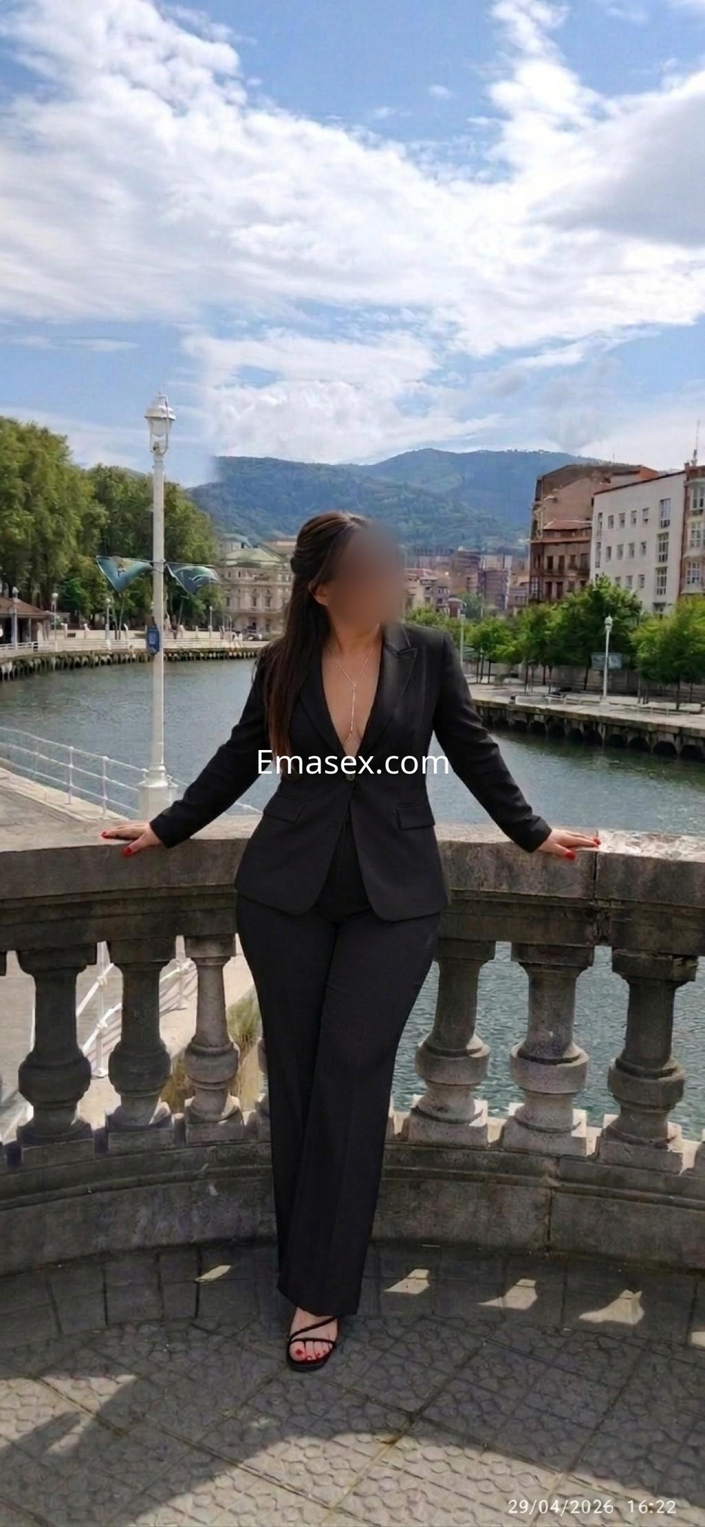 Escort de Bilbao se encuentra en un puente con vistas al río, vestida con un elegante traje negro y sandalias.
