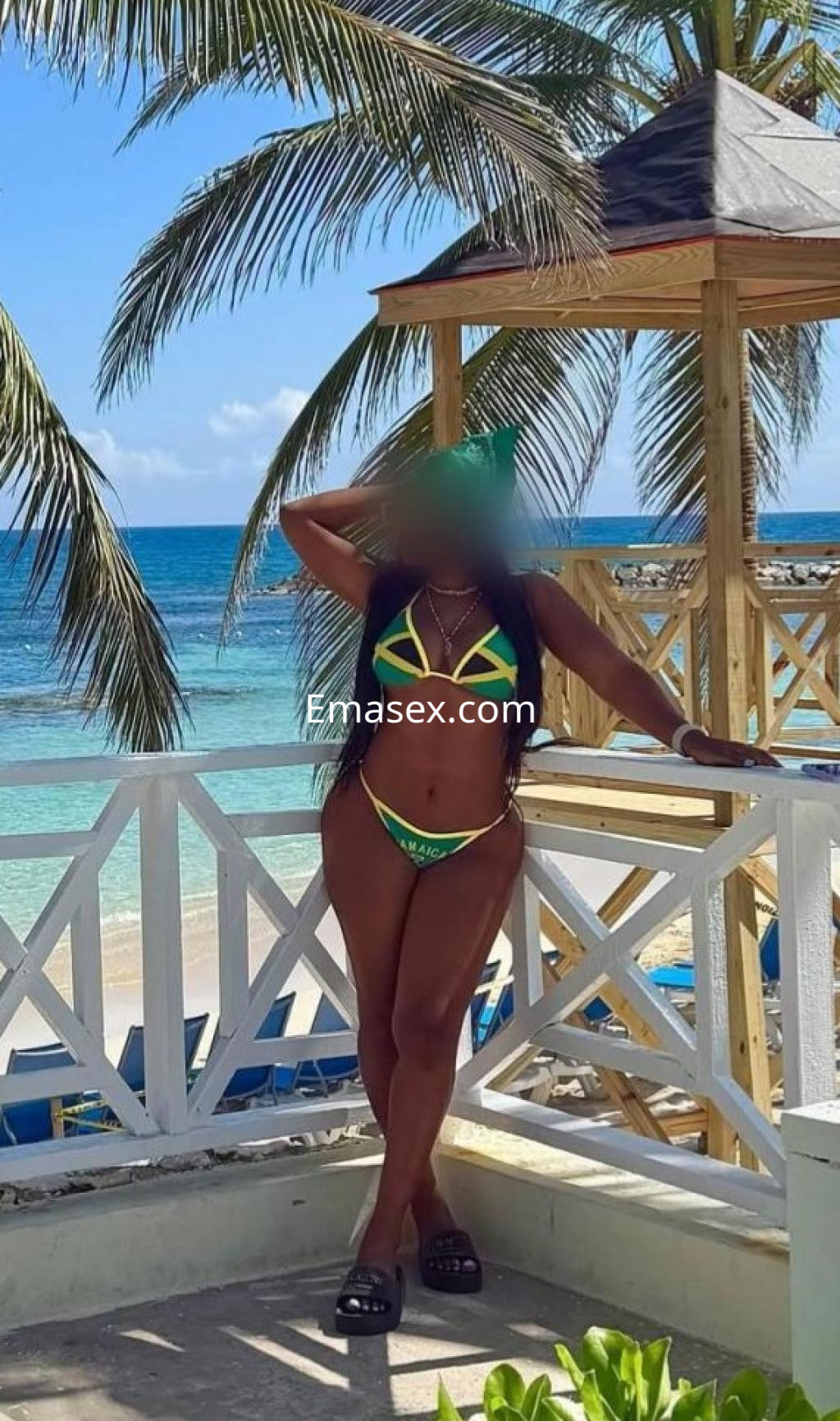 Escort en Albacete con bikini amarillo y verde, posando junto a una estructura de madera frente al mar.