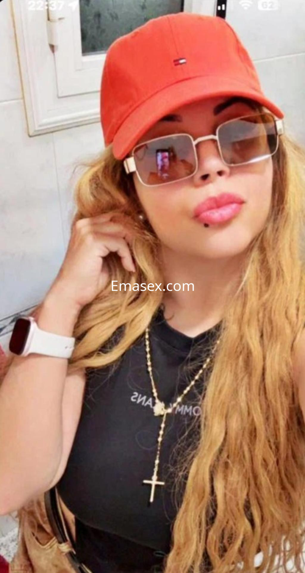 Escort en Barcelona con cabello largo y rizado, usando una gorra roja, gafas de sol y un top negro ajustado.