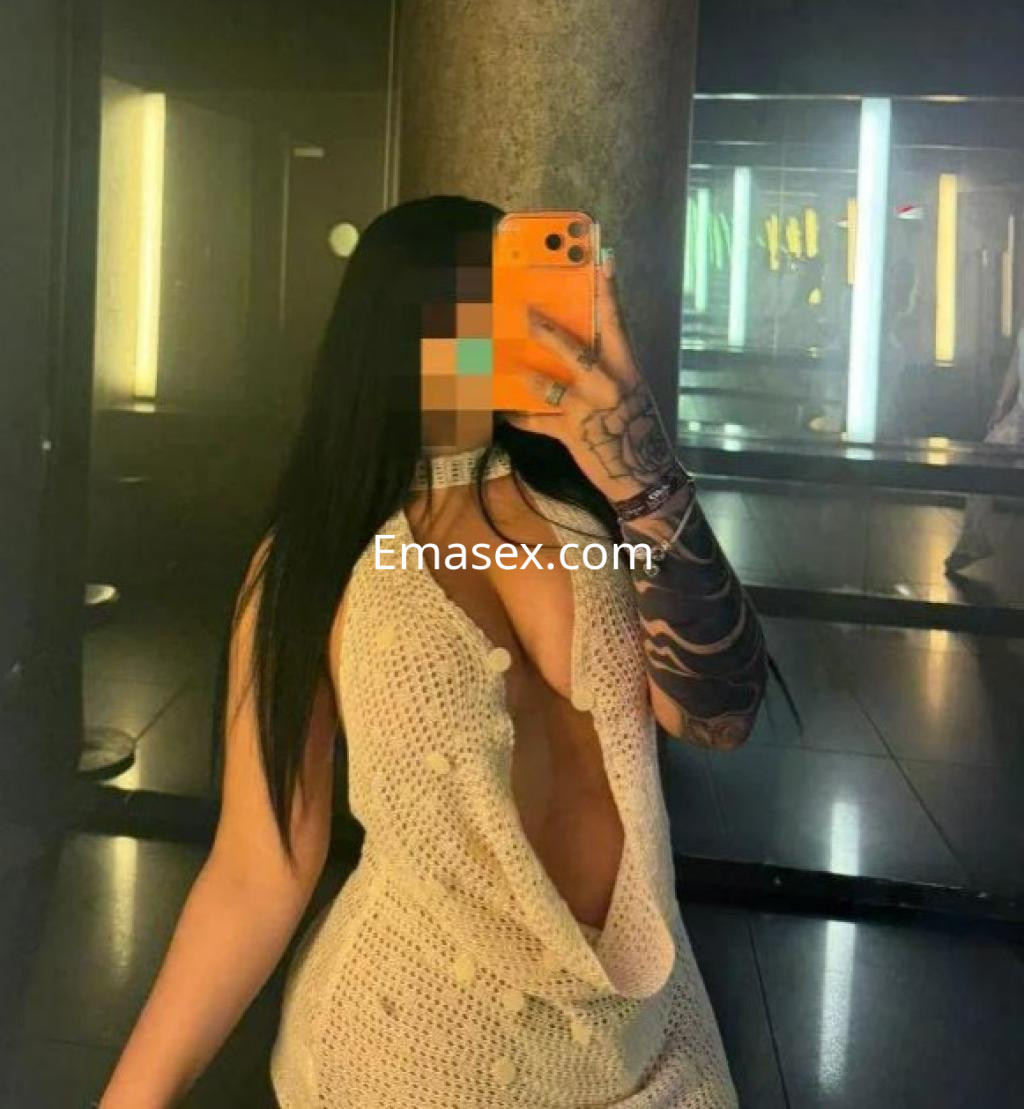 La puta en Barcelona se muestra con un vestido de punto abierto, posando frente a un espejo con un fondo oscuro.