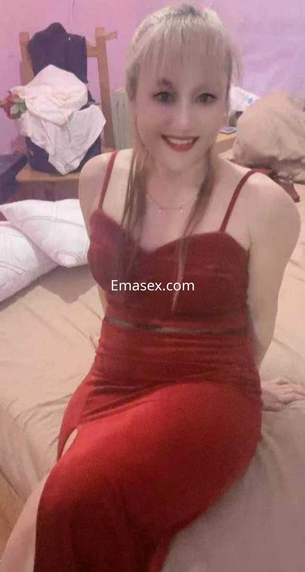 Puta sentada en una cama con un vestido rojo ajustado, sonriendo, en un ambiente acogedor en Barcelona.
