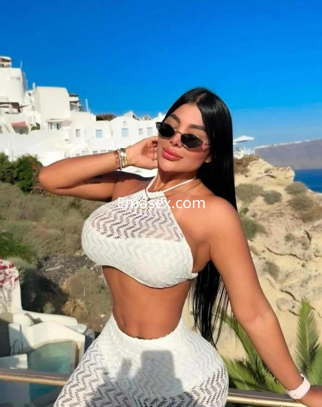 Puta en Alicante viste un conjunto blanco de crochet, posando con gafas de sol y un paisaje costero de fondo.