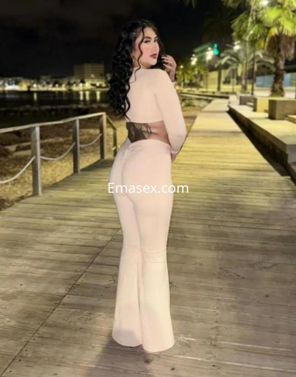 Escort posando de espaldas en un paseo marítimo de Alicante, luciendo un conjunto ajustado y cabello rizado.