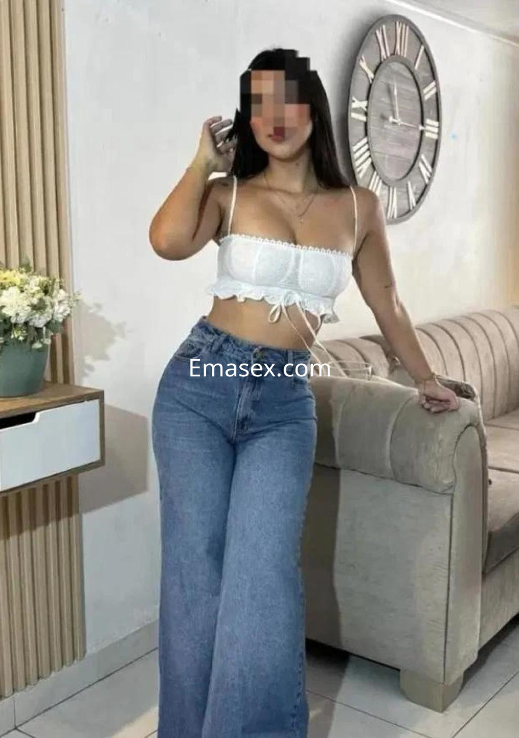 Escort en Zamora, luciendo un top blanco con tirantes y jeans de corte amplio, posando en un entorno acogedor.