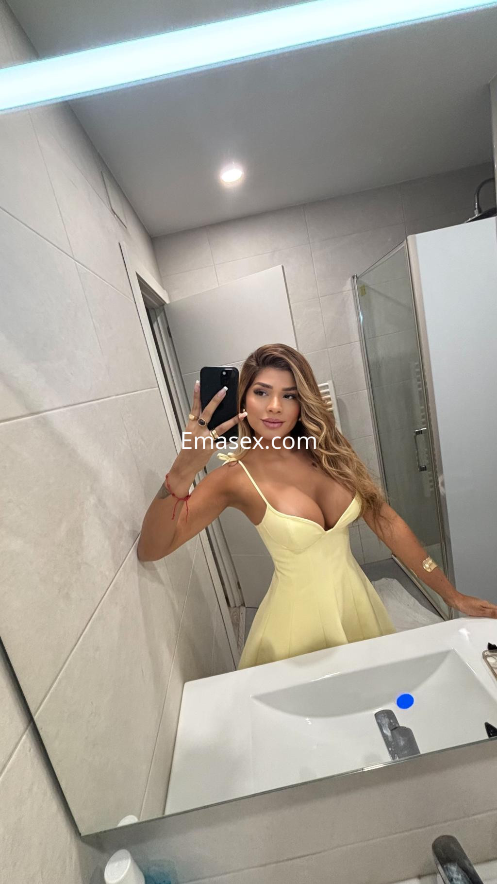 Puta en Bilbao posando frente a un espejo con un vestido amarillo, cabello largo y ondulado, en un baño moderno.