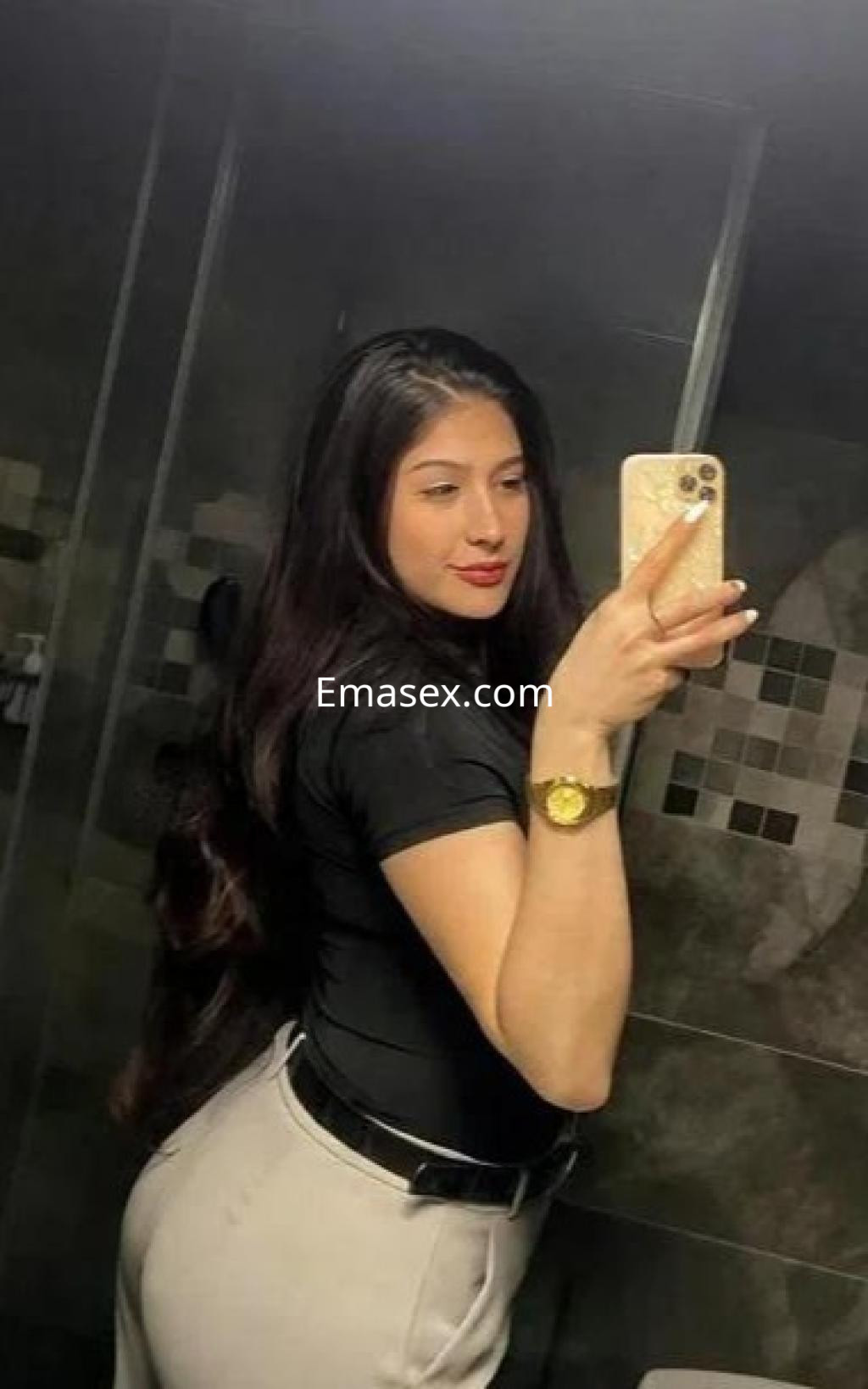 Puta con cabello largo y oscuro, de pie en un baño, usando una camiseta negra y pantalones claros, en Huelva.
