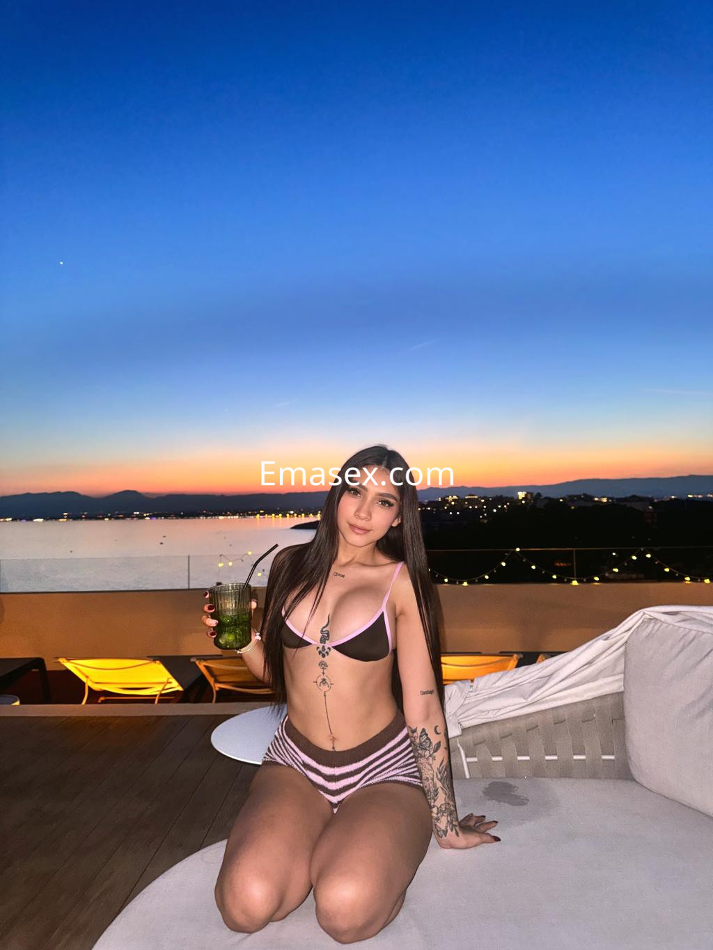 La puta se encuentra en una terraza con vistas al atardecer, vistiendo un bikini y sosteniendo una bebida.