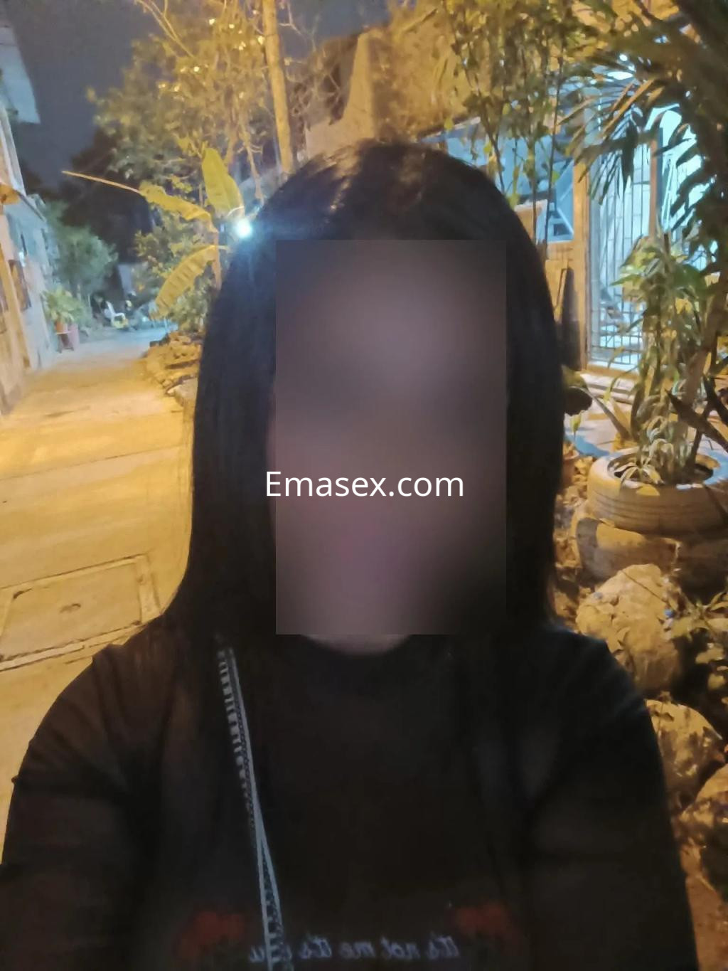 Escort en Albacete con camiseta negra y cabello largo, posando en una calle iluminada por la noche.