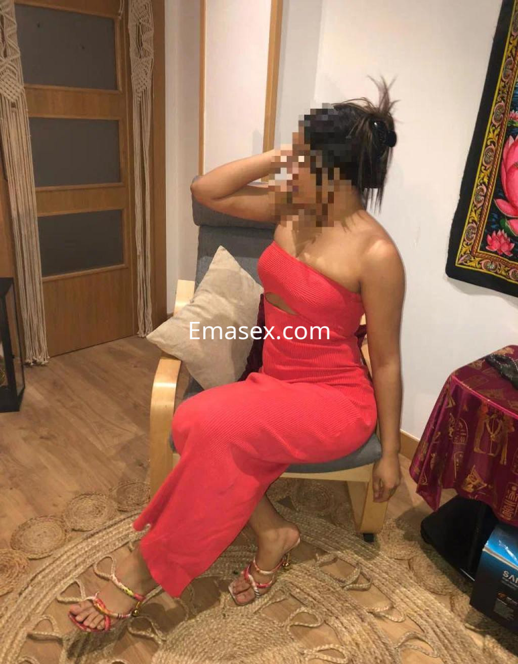 Escort en Madrid sentada en un sillón, vistiendo un vestido rojo ajustado y sandalias, con cabello recogido.