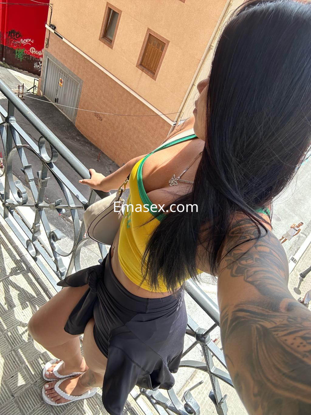Escort en Irún con cabello largo y oscuro, vistiendo un top amarillo y una falda negra, posando en un balcón.