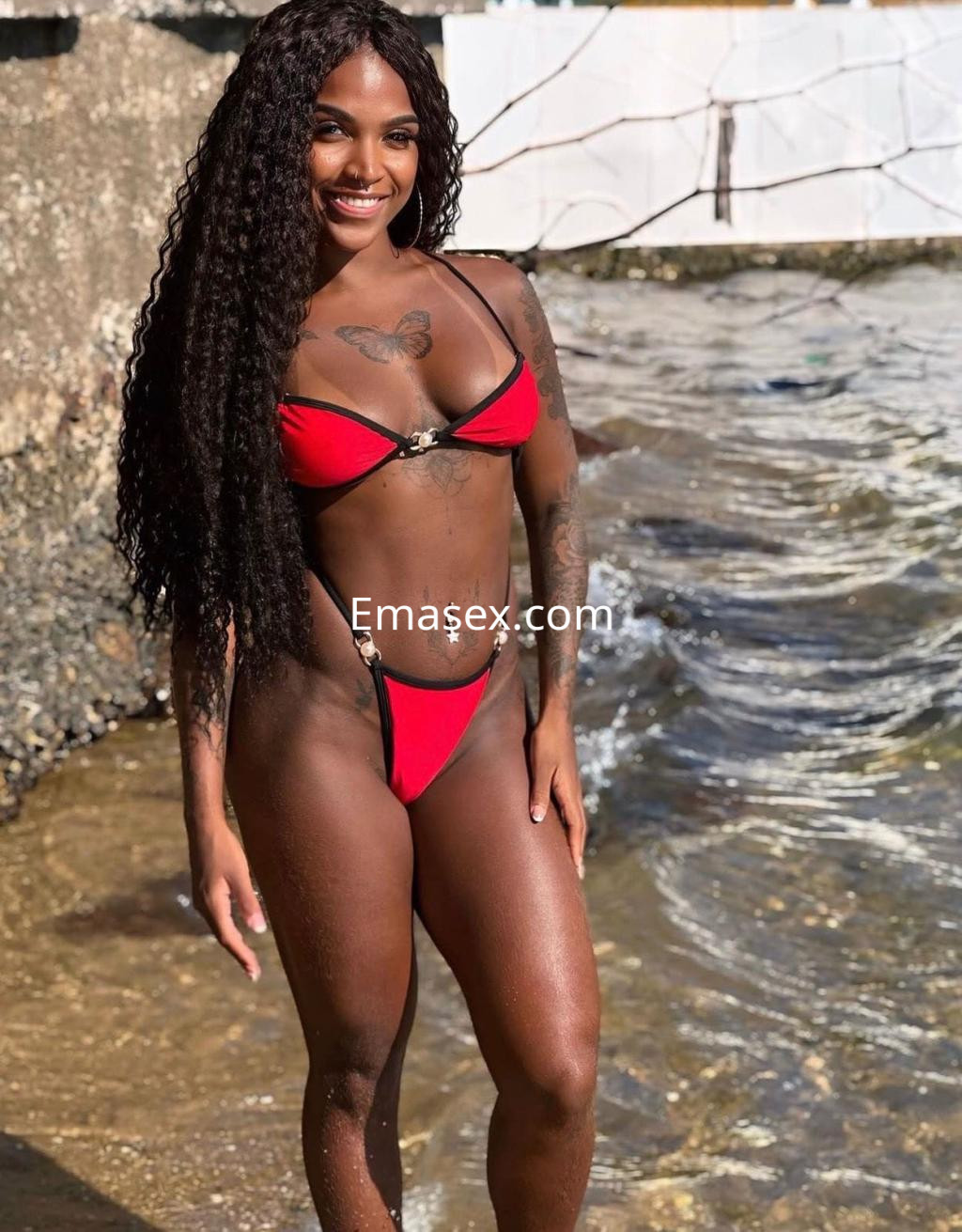 Puta en Bilbao con cabello largo y rizado, vistiendo un bikini rojo mientras posa junto al agua.