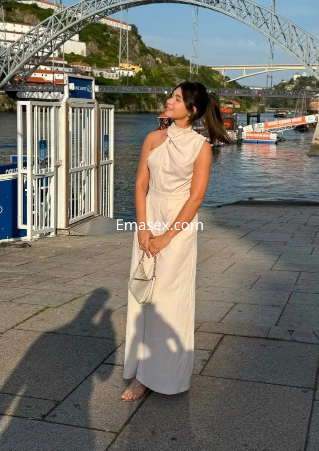 Escort en Palma de Mallorca, posando al aire libre con un vestido claro y una bolsa pequeña, frente a un río.