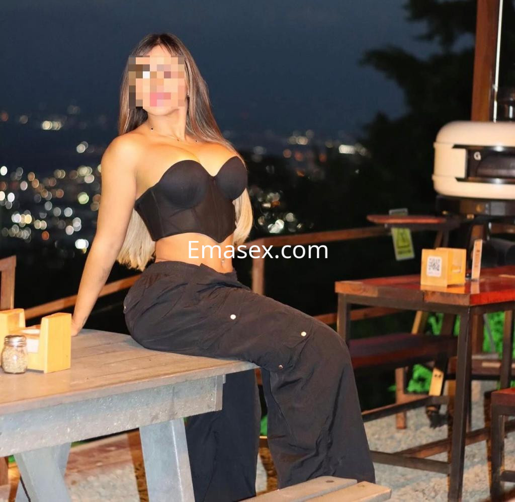 Puta posando en una terraza de Palma de Mallorca, con top negro y pantalones anchos, iluminada por la noche.