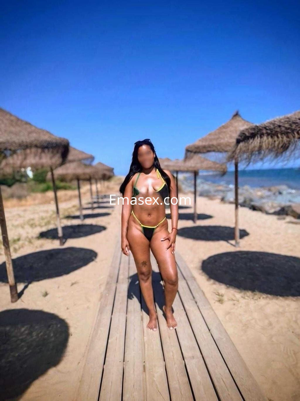 Escort en Huelva, posando en la playa con un bikini verde, junto a sombrillas de paja y mar al fondo.