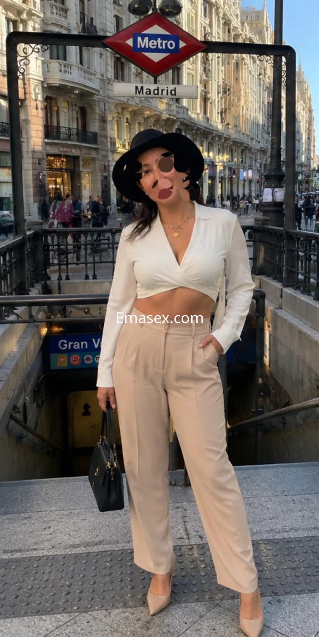 Escort en Murcia posando con una blusa corta blanca y pantalones claros, frente a una estación de metro.