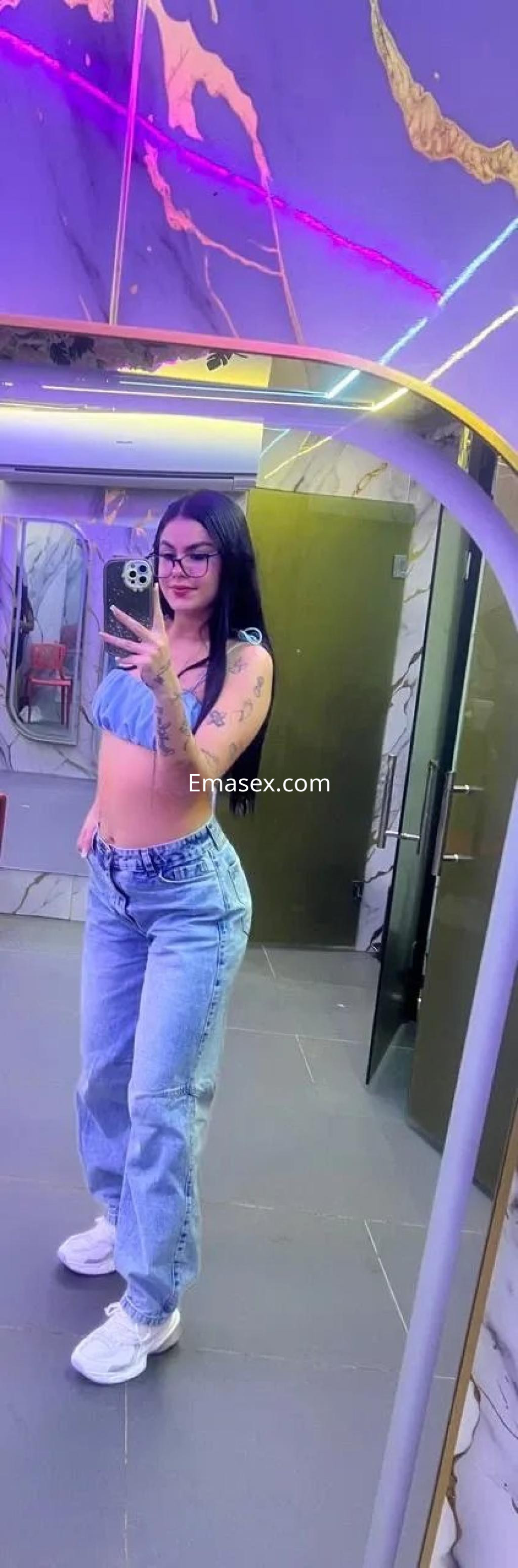 Escort en Oviedo posando frente al espejo, con top azul, jeans claros y zapatillas blancas en un ambiente moderno.