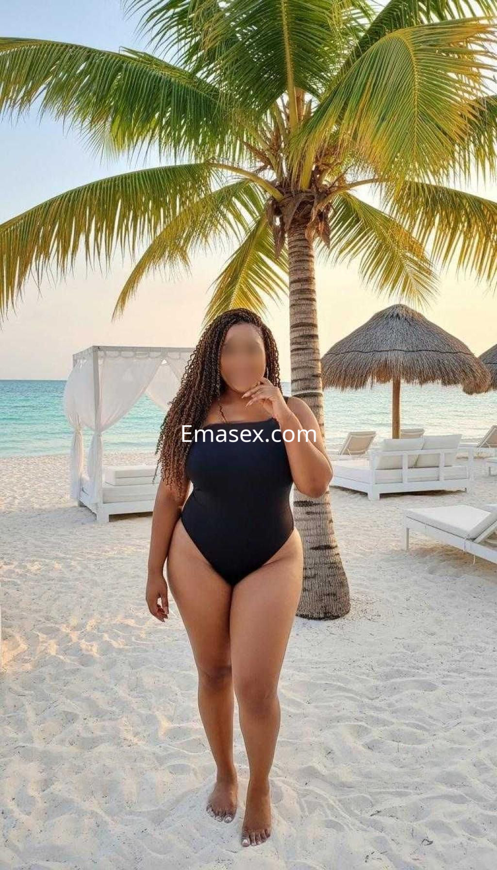 Puta con traje de baño negro posando en la playa de Tarragona, rodeada de palmeras y un ambiente tropical.