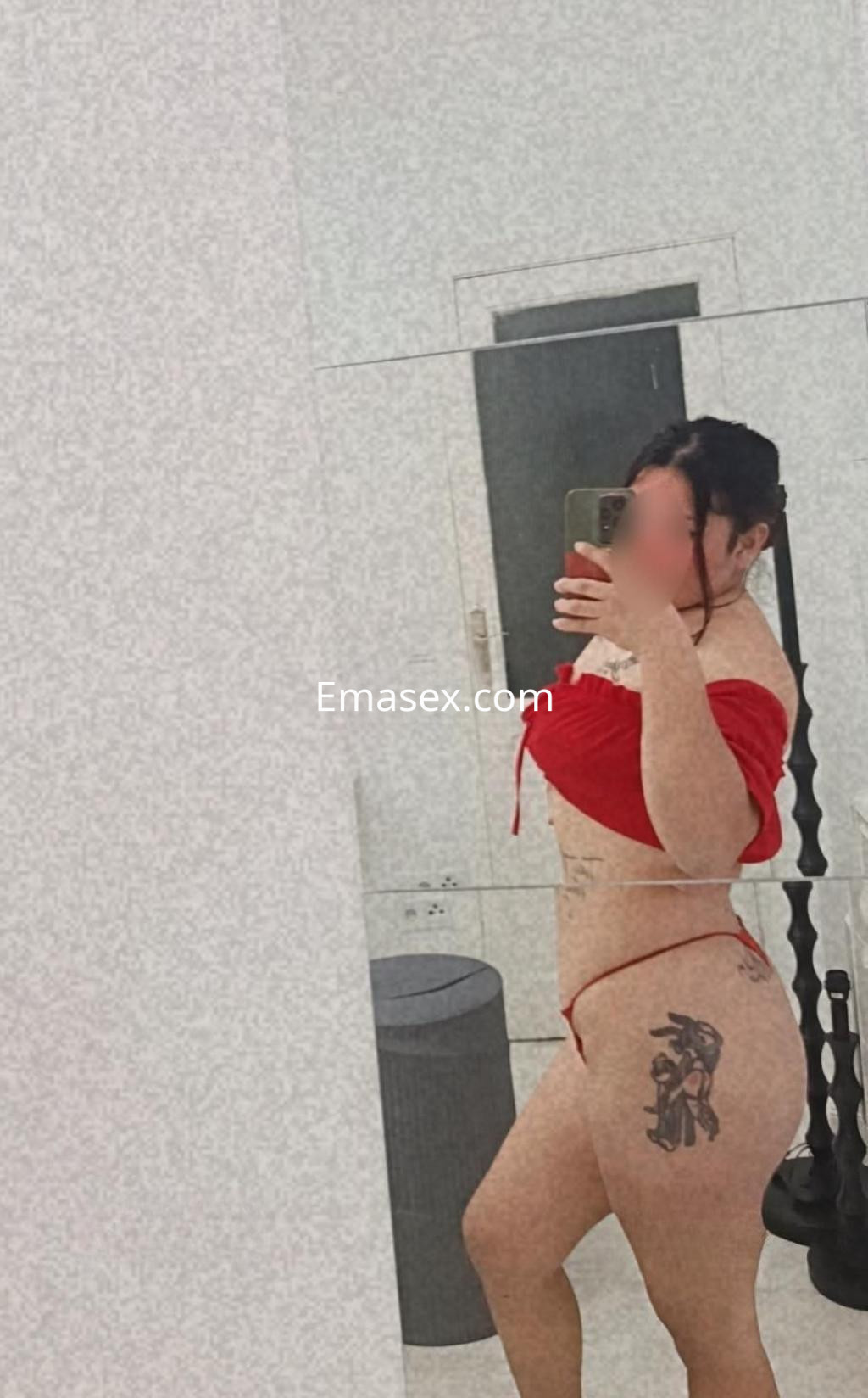 La puta en Tarragona luce un top rojo y una tanga, mostrando un tatuaje en su muslo en un entorno moderno.